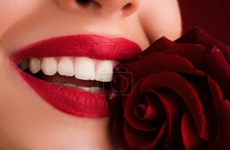 Foto de Labios con lápiz labial de cerca. Hermosos labios de mujer con sonrisa. Primeros planos hermosos labios femeninos con maquillaje rojo brillante. Piel limpia perfecta, maquillaje de labios sexy. Hermoso retrato de spa con tierna rosa roja - Imagen libre de derechos