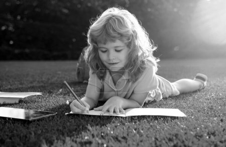 Téléchargez les photos : Garçon mignon avec écriture au crayon sur un cahier posé sur de l'herbe. Enfant lisant un livre dans le parc d'été - en image libre de droit