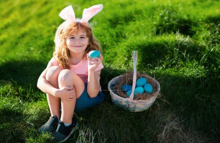 Téléchargez les photos : Joyeuses Pâques pour les enfants. Garçon dans les oreilles de lapin avec des œufs colorés jouer et chasser des œufs de Pâques à l'extérieur. Enfant mignon avec panier de Pâques sur l'herbe - en image libre de droit