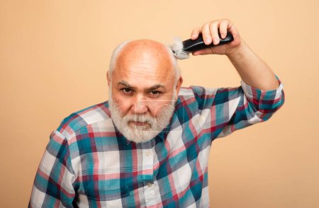 Téléchargez les photos : Un barbu se coupe les cheveux avec une tondeuse. Homme chauve d'âge moyen tondeuse à cheveux, Calvitie mature et concept de perte de cheveux - en image libre de droit