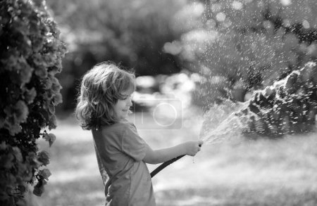 Foto de Divertido niño jugando con manguera de jardín en el patio trasero. Niño divirtiéndose con spray de agua en el fondo de la naturaleza patio. Actividades al aire libre para niños de verano - Imagen libre de derechos