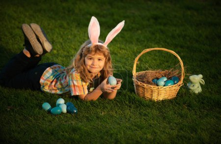 Téléchargez les photos : Des lapins avec des oreilles de lapin. Des enfants chassant des œufs de Pâques. Un gamin allongé sur l'herbe et trouvant des œufs de Pâques. Enfant avec œufs de Pâques et oreilles de lapin, portrait en plein air - en image libre de droit