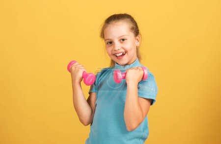 Foto de Cute little girl doing exercises with dumbbells in yellow background. Closeup portrait of sporty child with dumbbells. Happy child girl exercising - Imagen libre de derechos
