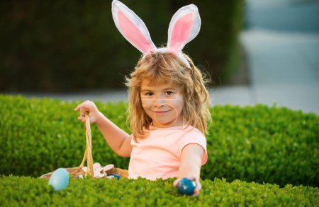 Foto de Niño cazando huevos de Pascua. Niño de Pascua en orejas de conejo cazando huevos de Pascua al aire libre. Lindo niño en traje de conejo con orejas de conejo divertirse en el parque - Imagen libre de derechos