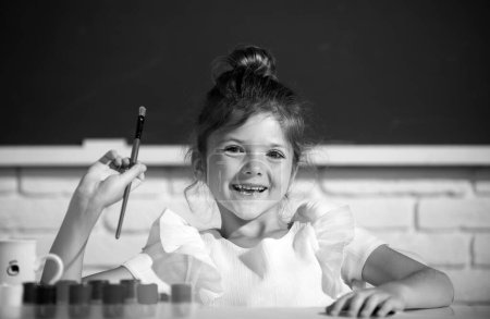 Foto de Child girl draws in classroom sitting at a table, having fun at school blackboard background. Painting school lesson, drawing art - Imagen libre de derechos