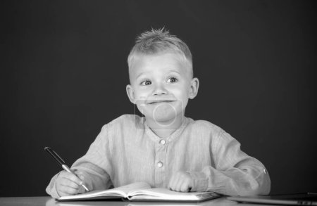Photo for Funny kid writing in notebook in class. Child at school. Kid is learning in class on background of blackboard - Royalty Free Image