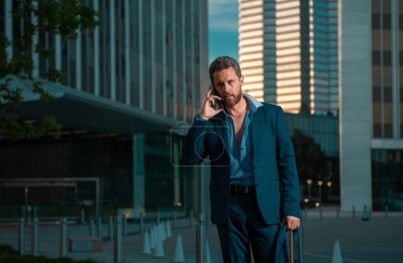 Téléchargez les photos : Portrait homme d'affaires sexy utilisant smartphone près du bâtiment moderne en plein air. Homme d'affaires prospère marchant en costume dans la rue - en image libre de droit