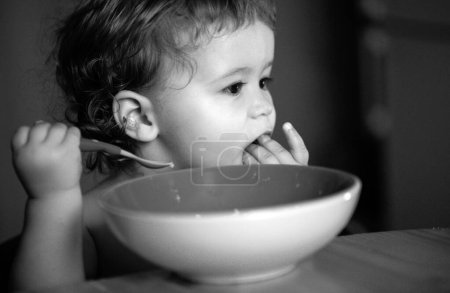 Téléchargez les photos : Bébé drôle manger de la nourriture lui-même avec une cuillère sur la cuisine - en image libre de droit