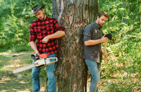 Foto de Dos amigos leñadores en el bosque. Cortador de madera con hacha y leñador con motosierra. Guapo leñador hombres hipsters en el bosque - Imagen libre de derechos