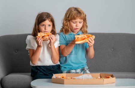 Foto de Niños hambrientos comiendo pizza. Niños pequeños amigos, chico y chica muerden pizza. Comida rápida poco saludable - Imagen libre de derechos