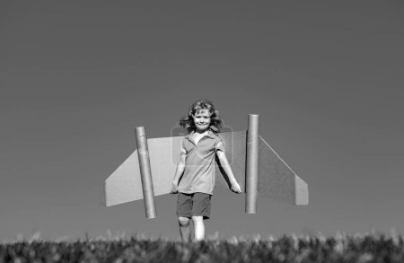 Happy child play with toy plane cardboard wings against blue sky. Kid having fun in summer field outdoor. Portrait of boy with paper wings