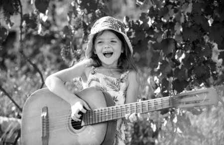 Téléchargez les photos : Child musician playing guitar. Stylish little child girl wearing a summer dress having fun on backyard. Dreamy kids face - en image libre de droit