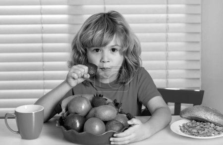 Téléchargez les photos : L'enfant mange des fraises, des fruits biologiques. Enfant préadolescent garçon dans la cuisine à la table manger des légumes et des fruits pendant le dîner déjeuner. Alimentation saine, plat de légumes pour enfants - en image libre de droit