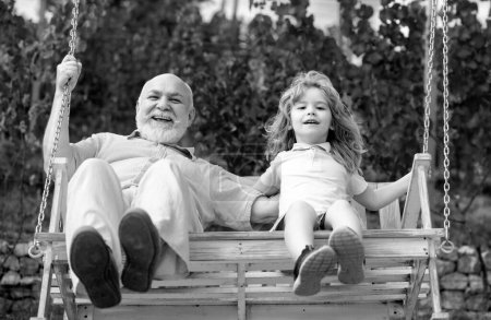 Foto de Emocionado abuelo asombrado y nieto niño pequeño balanceándose en el jardín al aire libre. Abuelo y nieto sentados en el columpio en el parque - Imagen libre de derechos