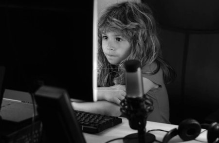 Téléchargez les photos : Petit gamer enfant jouer jeu d'ordinateur sur le bureau. Flux de jeu et blogging pour les enfants. École, étude, concept d'apprentissage en ligne - en image libre de droit