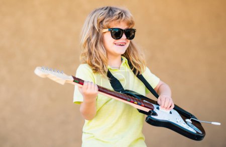 Foto de Gracioso músico hipster niño tocando la guitarra. Retrato de un niño divertido con gafas practicando una canción durante una lección de guitarra en la calle. Concepto musical, escuela de música para niños. Concierto de rock - Imagen libre de derechos