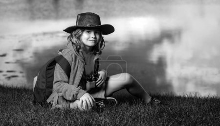 Foto de Lindo niño rubio con prismáticos con sombrero explorador y mochila en la naturaleza. Niño explorador senderismo y aventura - Imagen libre de derechos