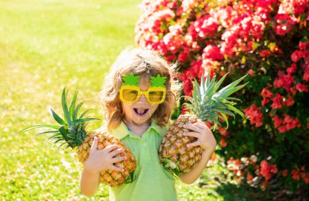 Téléchargez les photos : Joyeux petit garçon avec t-shirt tenant un ananas. Enfants avec fruits tropicaux en plein air - en image libre de droit