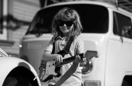 Téléchargez les photos : Enfant musicien jouant de la guitare comme un rockstar en plein air. Petit garçon musicien rock avec guitare. Portrait d'un enfant drôle avec des lunettes pratiquant une chanson lors d'un cours de guitare dans la rue. Concert rock - en image libre de droit