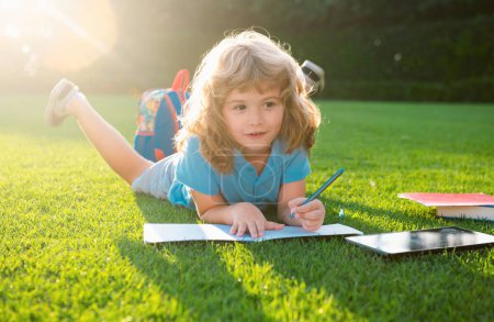Téléchargez les photos : Charmant enfant mignon avec écriture au crayon sur ordinateur portable à l'extérieur. Livre de lecture pour enfants dans le parc - en image libre de droit