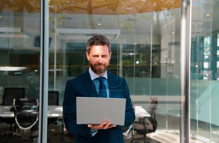 Téléchargez les photos : Homme d'affaires tenant un ordinateur portable debout dans la rue près de l'immeuble de bureaux d'affaires - en image libre de droit