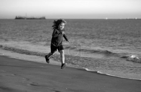 Foto de Niños jugando en la playa. Los niños juegan en el mar en las vacaciones familiares de verano. Arena y agua diversión, protección solar. Pequeño niño corriendo y saltando en la orilla del océano. Pequeño corredor haciendo ejercicio - Imagen libre de derechos