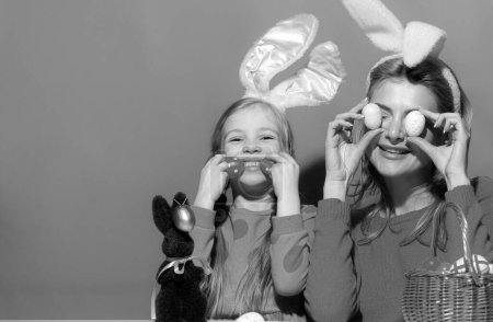 Foto de Retrato de madre con hija con orejas de conejo decorando huevos de Pascua aislados en azul - Imagen libre de derechos