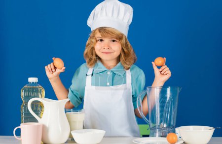 Téléchargez les photos : Portrait d'un enfant de 7, 8 ans coiffé d'une casquette et d'un tablier contenant des œufs qui font de la salade de fruits et cuisinent des aliments dans la cuisine. Mignon petit chef souriant heureux blonde - en image libre de droit