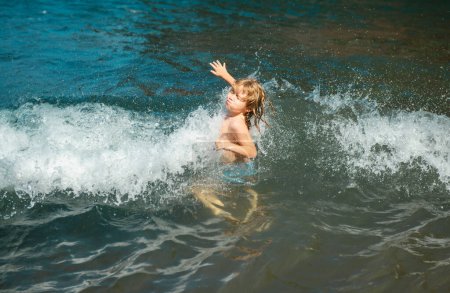Téléchargez les photos : Happy kid amusez-vous sur la plage tropicale de la mer. Drôle de garçon avec des éclaboussures par l'eau. Mode de vie actif des enfants, activité de natation pendant les vacances d'été avec les enfants - en image libre de droit