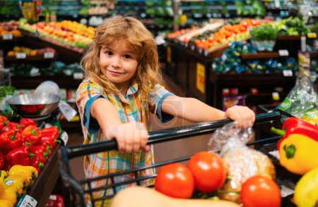 Foto de Child in supermarket buying fruit. Kid grocery shopping. Kid with cart choosing fresh vegetables in local store. Healthy kids food. Child in supermarket buys vegetables - Imagen libre de derechos