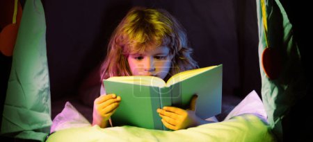 Foto de Child reading a book in the dark home. Close up portrait of Boy sitting on in living room watching pictures in story book. Kid doing homework for elementary school. Children study - Imagen libre de derechos