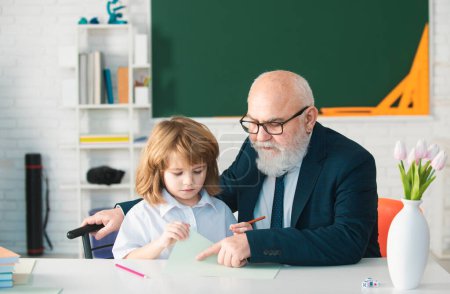 Photo for Elementary pupil reading and writing with teacher in classroom. Old senior teacher and school boy pupil - Royalty Free Image