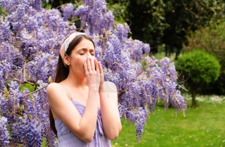 Girl sneezing with allergy symptom concept. Woman being allergic to blossom during spring front of blooming tree outside