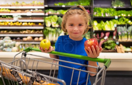 Téléchargez les photos : Enfant avec pomme et caddie à l'épicerie. Drôle mignon enfant sur le shopping au supermarché. L'épicerie. Magasins d'alimentation, mode de vie sain concept - en image libre de droit