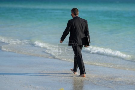 Foto de Vista trasera del hombre de negocios espalda en traje en agua de mar en la playa. Trabajo remoto en línea. Loco negocio de verano. Divertido estilo de vida empresarial. Divertido empresario freelance - Imagen libre de derechos