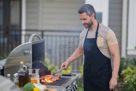 Foto de Hombre hispano cocinando en la barbacoa en el patio trasero. Chef preparando barbacoa. Maestro del chef de barbacoa. Hombre guapo preparando barbacoa, carne de barbacoa. Parrilla y barbacoa - Imagen libre de derechos