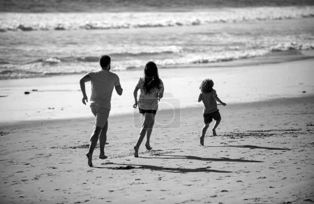 Téléchargez les photos : Joyeux jeune famille courir et sauter sur la plage d'été. Enfant dont les parents courent et sautent. Concept de famille amicale et de vacances d'été - en image libre de droit