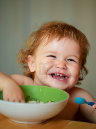 Photo for Funny baby eating food himself with a spoon on kitchen. Smiling baby eating food - Royalty Free Image