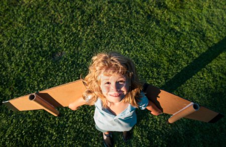 Téléchargez les photos : Enfant garçon rêve et voyage sur l'herbe verte dans le parc. Garçon avec jouet d'avion à l'extérieur. Enfant heureux jouant avec jouet avion à l'extérieur dans le champ d'été. Voyage, vacances et concept de liberté - en image libre de droit