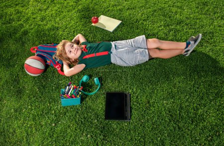 Téléchargez les photos : Vue du dessus d'un élève d'âge préscolaire allongé sur l'herbe et prenant un cours à l'extérieur. Garçon heureux relaxant sur l'herbe - en image libre de droit