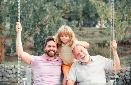 Photo for Boy son with father and grandfather swinging together in park outdoors. Three different generations ages grandfather father and child. Dad, son and granddad hugging - Royalty Free Image