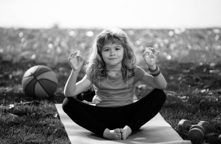 Téléchargez les photos : Petit enfant assis sur le tapis roulant pratiquant le yoga méditer dans le parc en plein air. Enfant garçon pratiquant la pose de yoga - en image libre de droit