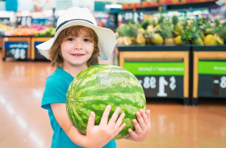 Téléchargez les photos : Enfant tenant pastèque au supermarché contre étal de nourriture. Magasinage garçon positif souriant au supermarché - en image libre de droit