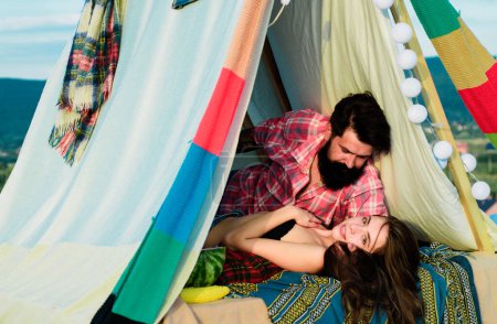 Téléchargez les photos : Couple romantique baiser en tente camping sur fond de nature. Aventure pour les jeunes amis le jour d'été. Couple hipster tendre baisers dans la tente du camp - en image libre de droit