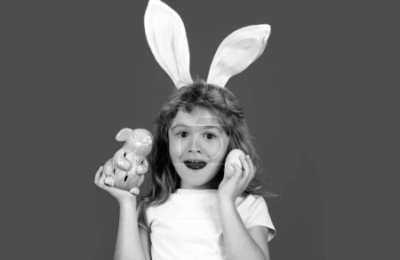 Foto de Niño excitado cazando huevos de Pascua. Retrato de asombrado, niño en traje de conejo con orejas de conejo teniendo pascua, aislado sobre fondo azul - Imagen libre de derechos