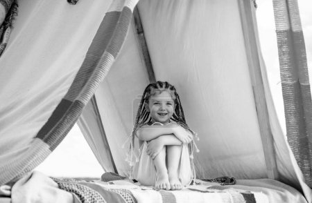 Téléchargez les photos : Un enfant heureux avec des dreadlocks joue dans une tente. Concept de camping pour enfants. Une fille qui joue au camp. S'amuser à l'extérieur au camping - en image libre de droit