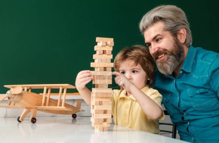 Foto de Sonriente padre e hijo enfocado jugando jenga juego en casa. Pequeño alumno con expresión de cara feliz cerca del escritorio con útiles escolares - Imagen libre de derechos