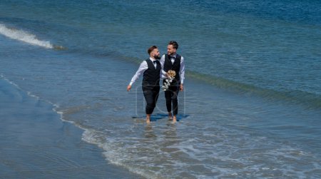 Foto de Los novios gay caminando juntos en la playa del mar durante el día de la boda. Hombres románticos en el agua de mar. Pareja homosexual celebrando su boda. Pareja LBGT en ceremonia de boda - Imagen libre de derechos