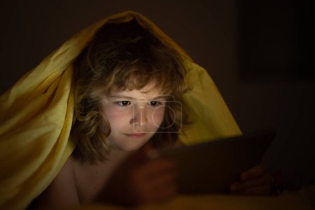 Téléchargez les photos : Enfant regardant la tablette avant de dormir. L'enfant lit l'e-book. Petit garçon regarde l'écran de la tablette à hauteur. Petit garçon jouant à la tablette allongé sur un lit sous une couverture. Addiction aux médias sociaux pour enfants - en image libre de droit