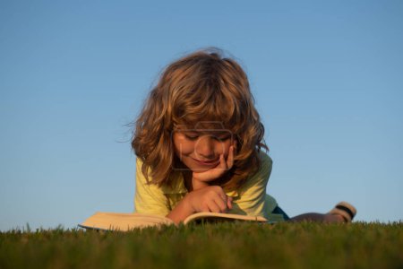 Téléchargez les photos : Enfant intelligent garçon lecture livre posé sur l'herbe sur l'herbe et le ciel de fond avec espace de copie - en image libre de droit